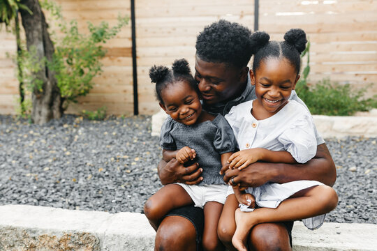 A father sitting outdoors in the backyard having a conversation with his two young daughters & giving them a bear hug! 