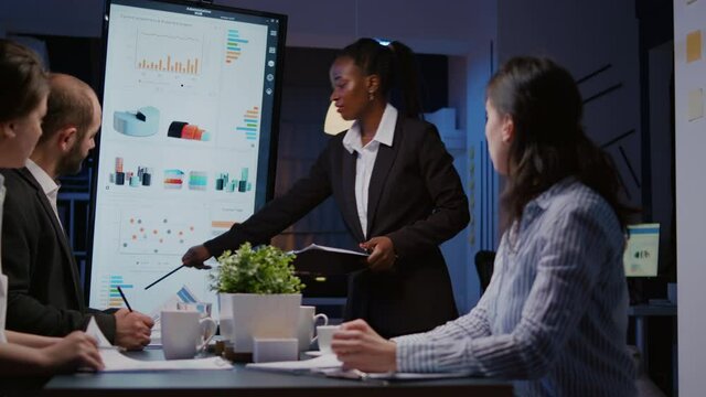 Businesswoman with dark skin explaining financial graphs using presentation monitor overworking in company meeting room in evening. Workaholics multi-ethnic teamwork solving statistics at night