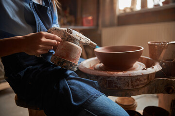 Beautiful elegant craft woman wearing apron making clay bowl on pottery wheel in workshop