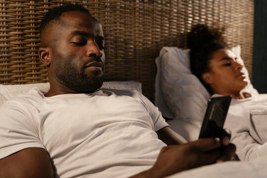Sad black couple lying in bed and browsing gadgets
