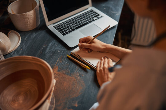 Unrecognized female artisan sitting at the work desk and looking to the screen of her laptop in pottery workshop - Powered by Adobe