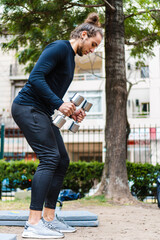 vertical photo of a young Brazilian long-haired man exercising biceps with metal weights in an outdoor city park.