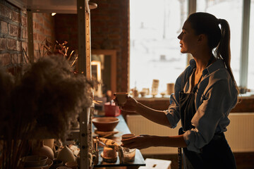 Beautiful Caucasian woman with clay tableware in hands standing near the shelf in art studio