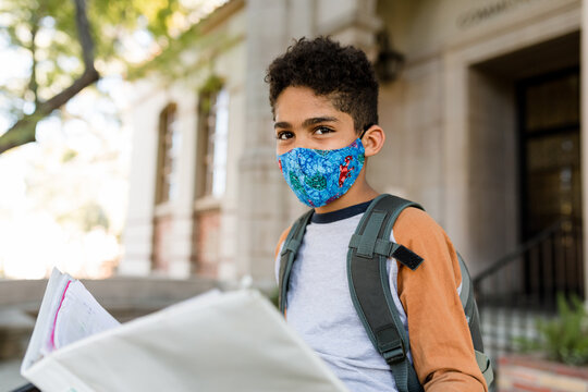 Boy Wearing a Mask Overlooks His Homework While Waiting on the Steps Outside School