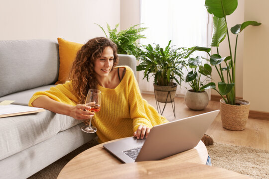 Smiling young woman drinking wine and watching video on laptop at home