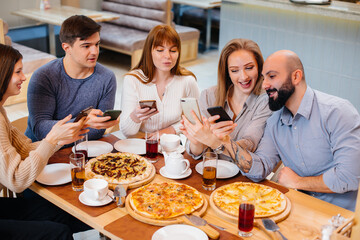 A group of young cheerful friends is sitting in a cafe talking and taking selfies on the phone. Lunch at the pizzeria.