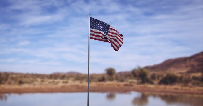 Composition of american flag on flagpole billowing over lake, landscape and blue cloudy sky