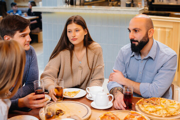 A group of young cheerful friends is sitting in a cafe talking and eating pizza. Lunch at the pizzeria.
