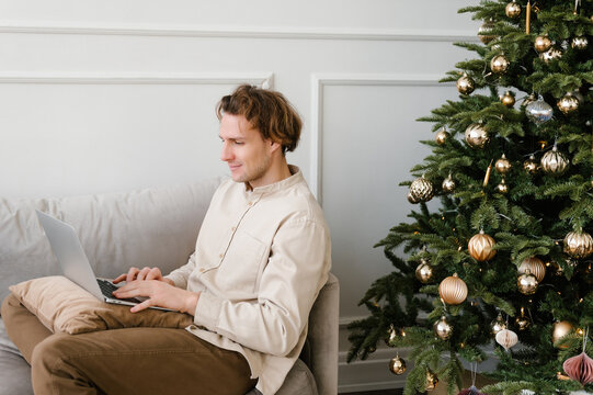 Serious young guy using laptop sitting on sofa near decorated Christmas tree at home