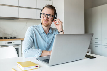 Smiling young male freelancer wearing a headset and looking away, a positive man with the beard working remotely in the customer service department, making and receiving calls