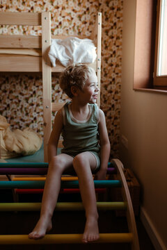 Blond child siting on a beautiful rainbow pikler in his room and looking out of the window