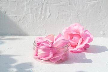  Beauty jar filled with natural rose petals standing next to rose flower on a white background with shadows from rose bush. rose oil natural beauty product mock up idea