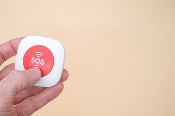 Elderly hand holding a wireless SOS emergency alarm button isolated on light brown background