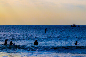 A silhouette of a man paddling a stand up paddle board in the sunrise lights with surfers looking at him. Beautiful golden and blue tones