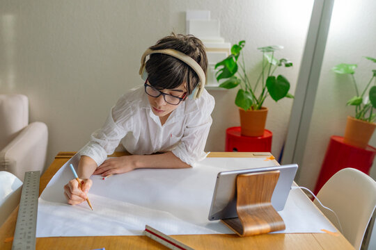 Pre-teen Boy wearing headphones drawing a map on big paper on a table with a device