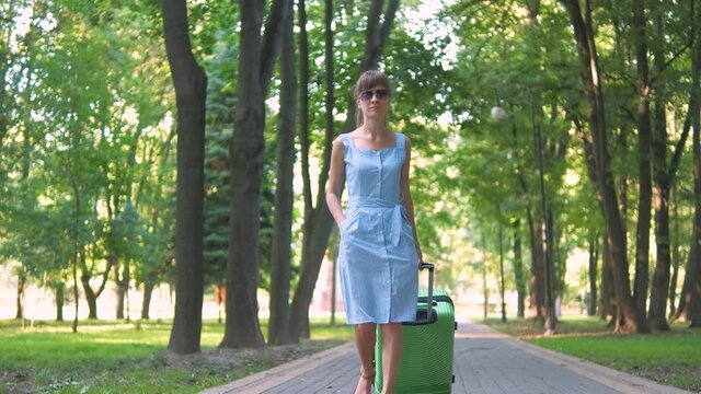 Young woman walking on park sidewalk with green suitcase on summer day. Travelling and vacation concept.