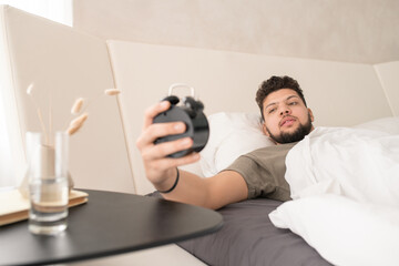 Young man looking at time on face of alarm clock in the morning