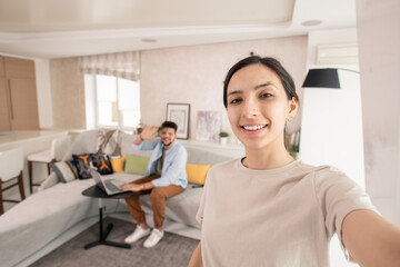 Happy female making selfie with her husband on background in home environment