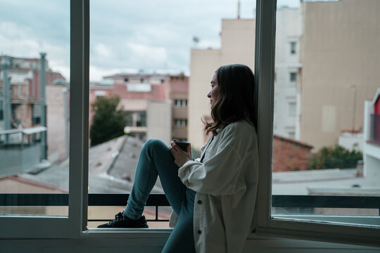 Young woman sitting on a window sill drinking warm tea