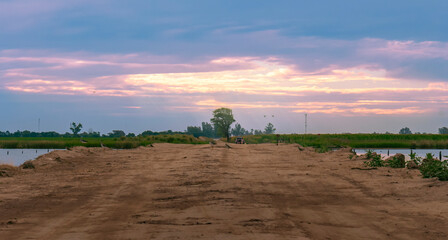 Obraz premium Scenic view of a sunset in a slightly cloudy sky on a rural field out of the city. View in perspective through a dirt road with an ancient tree in the background.