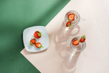 Infused water with strawberries in glasses and berries on a saucer on a green-pink background in hard light with shadows. Detox drink. Top view