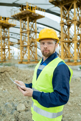 Serious builder in uniform and hardhat using tablet and looking at you