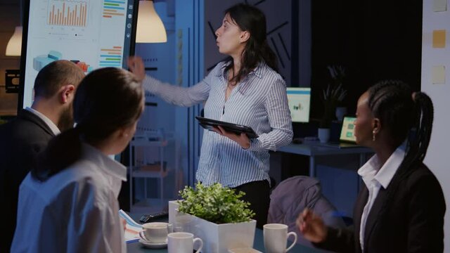 Workaholic focused businesswoman explaining management solution pointing strategy on monitor overworking in company business office meeting room. Multi-ethnic coworkers discussing ideas in evening