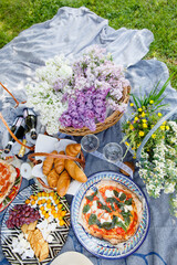 Wicker basket with lilacs, champagne glasses and pizza. Summer picnic in nature. Beautiful summer still life	
