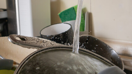 The kitchen utensils in the wash basin need to be washed. A pile of dirty dishes in the kitchen sink with running water. Kitchen utensils need washing. Homework concept.