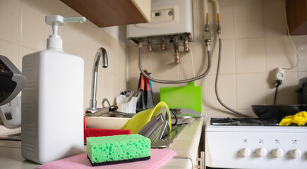 Green sponge and liquid soap dispenser for washing dishes on a dirty sink completely with dishes and kitchen utensils. Washing dishes in the kitchen by hand with detergent and a sponge.