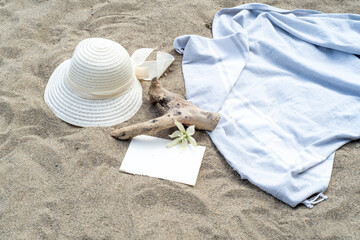 Summer still life. White straw hat on the sand of the mediterranean beach. Summer party invitation card mockup with white flower, branch and blue towel. Concept of vacation and relaxation. 