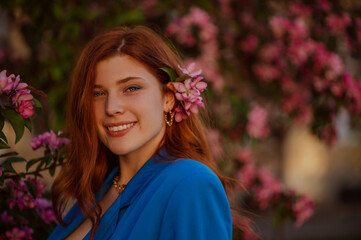 Outdoor close up portrait of happy smiling redhead freckled woman with blue eyeliner makeup posing near pink blooming tree. Copy, empty space for text
