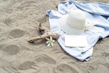 Summer still life. White straw hat on the sand of the mediterranean beach. Summer party invitation card mockup with white flower, branch and blue towel. Vacation and relaxation concept.