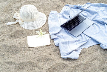 Summer still life. White straw hat on the sand of the mediterranean beach. Business card mockup with white flower, blue towel and laptop. Vacation and relaxation concept.