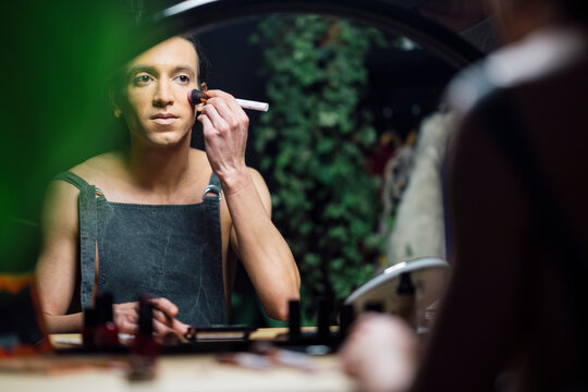 man sitting in front of mirror and touching face while doing makeup in drag queen style