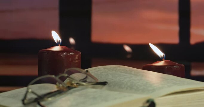 There are two burning candles on the table. In front of them is an open book, on which are reading glasses with diopters. Outside the window, the river, the forest at sunset.