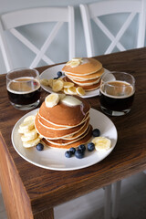 Stack of Tasty American style pancakes with a piece of butter, banana and blueberry on a white plate and two glasses of black American coffee. Breakfast for two or weekend brunch