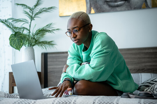 Young African woman working on her laptop while sitting on bed.