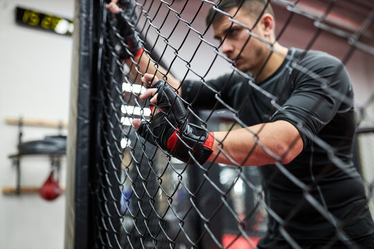 Brunette male in black wear and gloves practice kickboxing, mma sport concept, stand exhausted and tired after fighting. Side view portrait. Have rest, view through cage. focus on hands in gloves