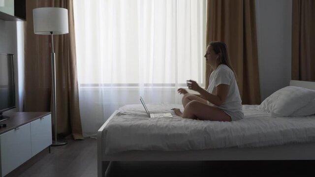 Woman in white T-shirt sits on bed near the window in headphones and works on notebook in the early morning alone at home in quarantine. Typing text on the keyboard on the computer