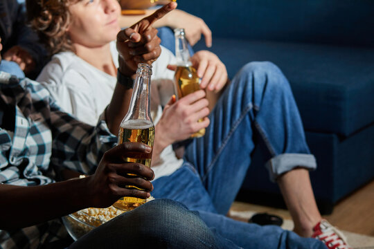 Interracial group of friends eating popcorn and drinking beer at home, enjoy watching movie. Friendly students gathered at night to spend time together, relaxed, dressed casually. focus on bottle