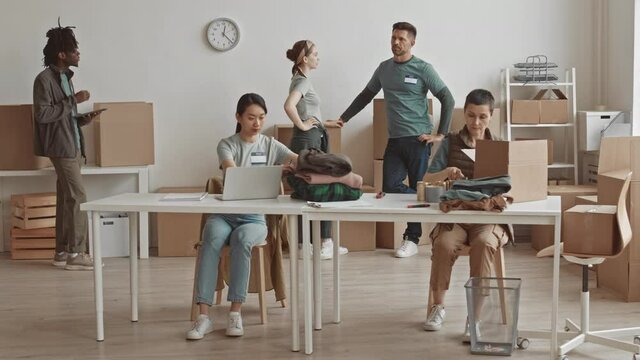 Full-shot slowmo of small team of multi-ethnic males and females working together at non-profit charity organization helping poor and needy people by packing clothes and food for them