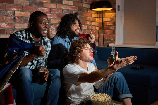 Group Of Diverse Men Sitting On Sofa Watching Sport Championship Together Emotionally Reacting On Game Process, Gesturing, Drinking Beer, At Home