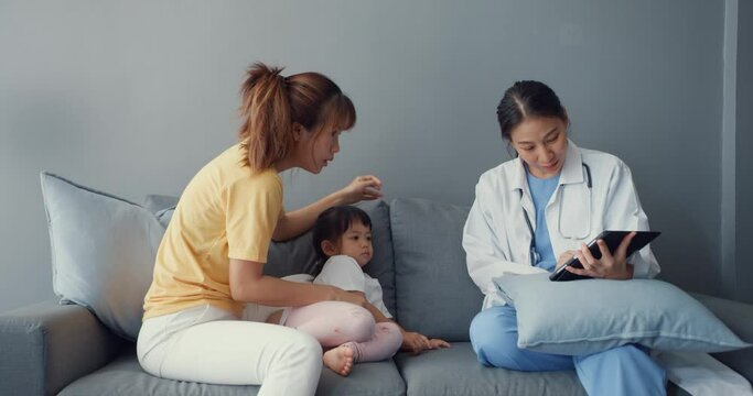 Young Asia female pediatrician doctor and little girl patient using digital tablet sharing good health test news with happy mom sit on couch in house. Medical insurance, Visit patient at home concept.
