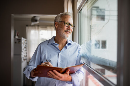 Older Gentleman Writing in a Notebook Looks Out His Apartment Window