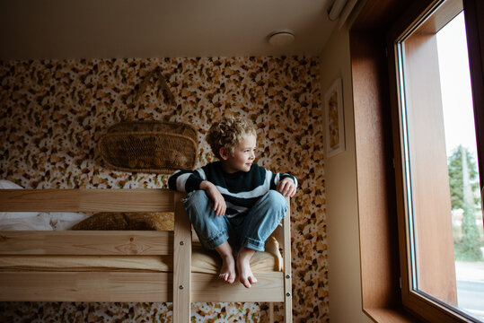 Toddler boy sitting on his wooden bunk bed and looking outdoors