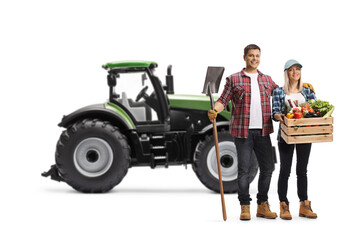 Full length portrait of a male and female farmers with a tractor holding a shovel and a crate with vegetables