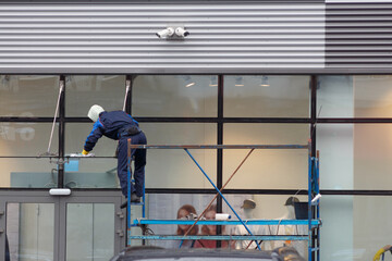 An employee of a cleaning company in blue overalls on a scaffold washes the glass facade of a store.