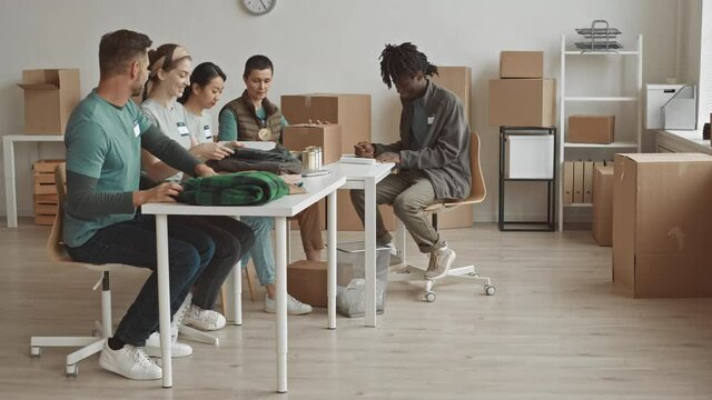 Full-shot slowmo of diverse volunteer team having conversation while sitting by table and packing donation boxes with clothes and eatables