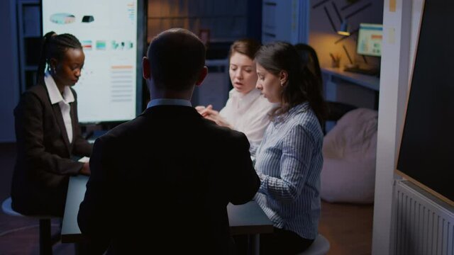 Selective focus of workaholics multi-ethnic businesspeople solving overtime company problem analyzing paperwork statistics. Focused coworkers overworking in business meeting room late at night